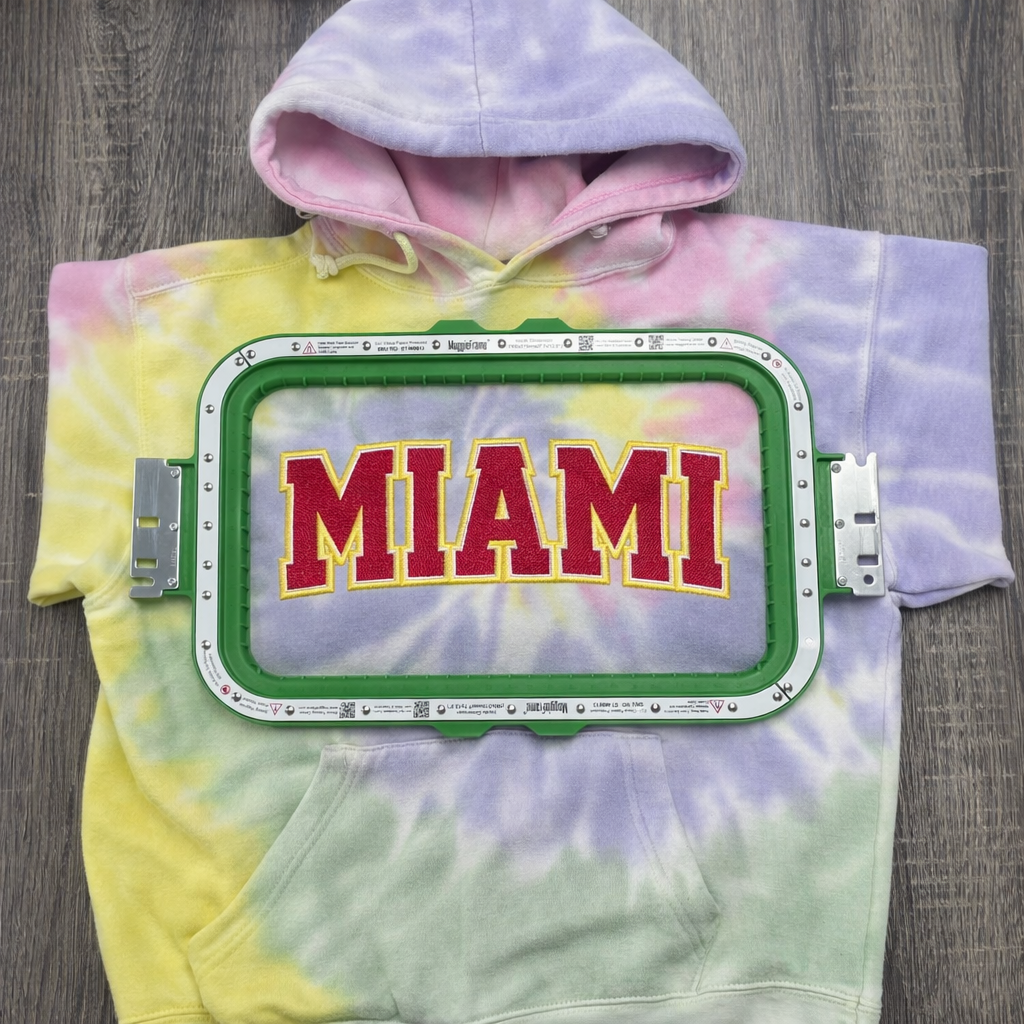 Tie-dye hoodie with 'MIAMI' text on a Green embroidery machine hoop with dimensions 7.7" x 12.5" on a white background