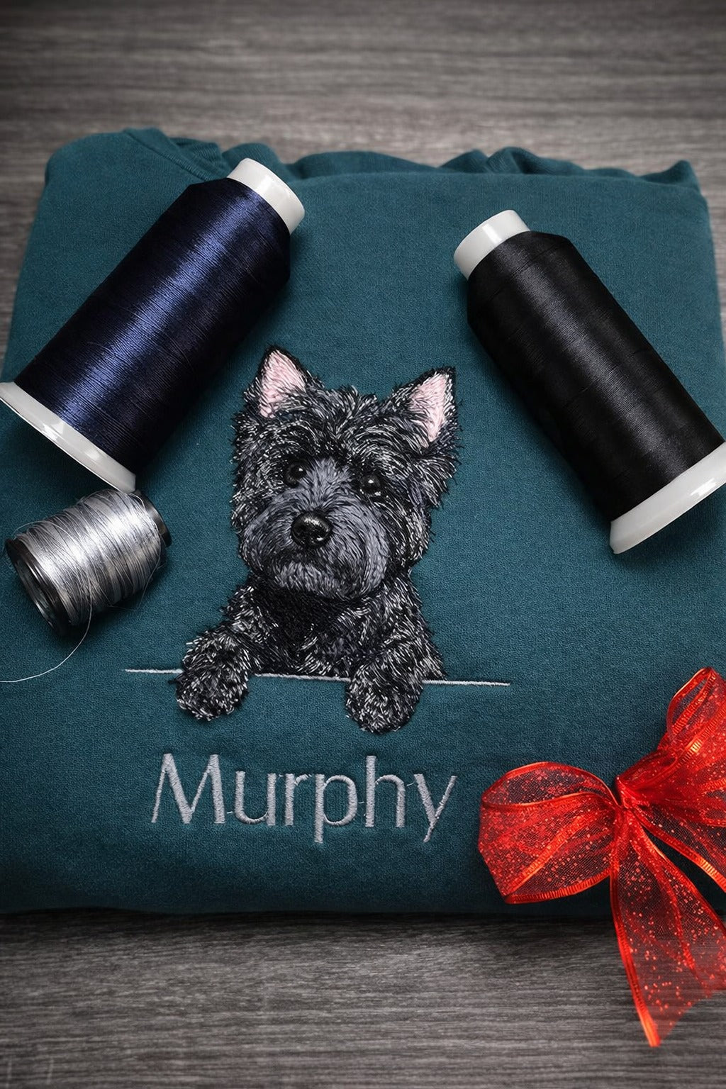 Teal pillow with embroidered dog and 'Murphy' text, surrounded by spools of thread and a red bow.
